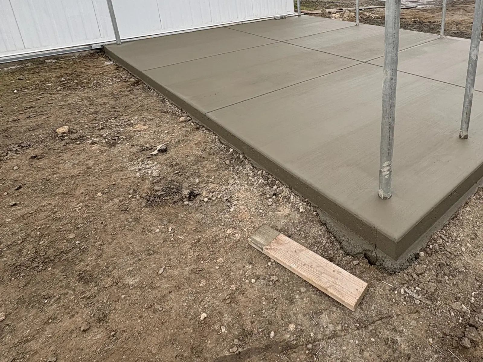 A new broom finished concrete pad with sawcut joints and steel posts set in it, poured beside a white metal building