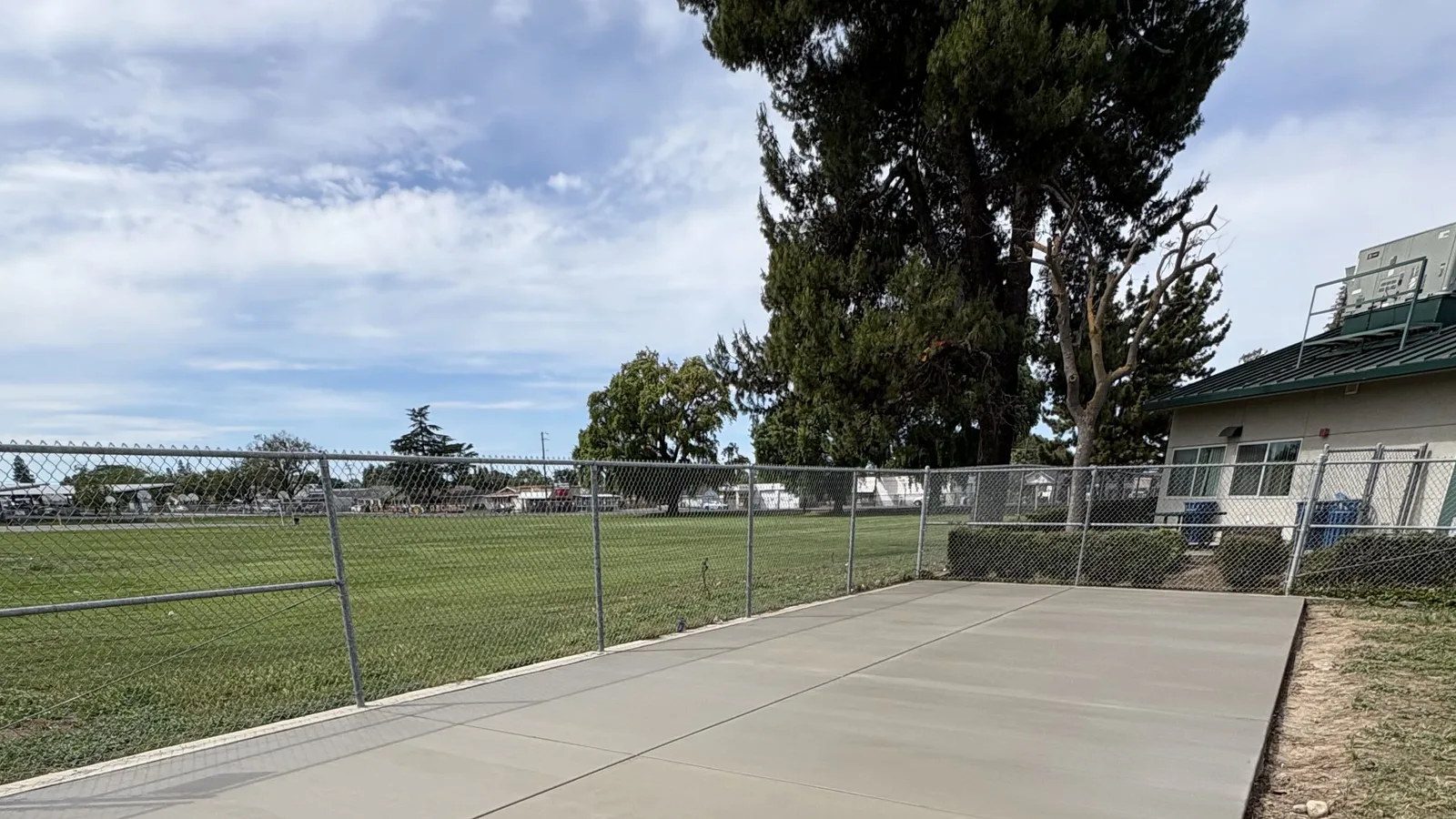 Wide view of concrete flatwork along a fence line at a sports field under a blue sky with clouds