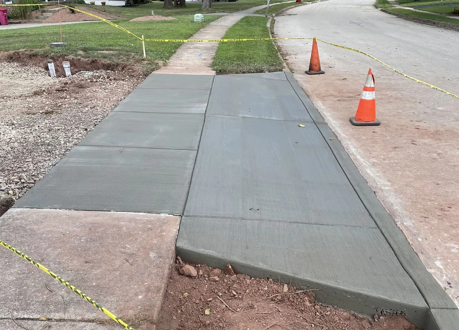 Freshly poured concrete driveway apron and sidewalk panels at the street edge, roped off with caution tape