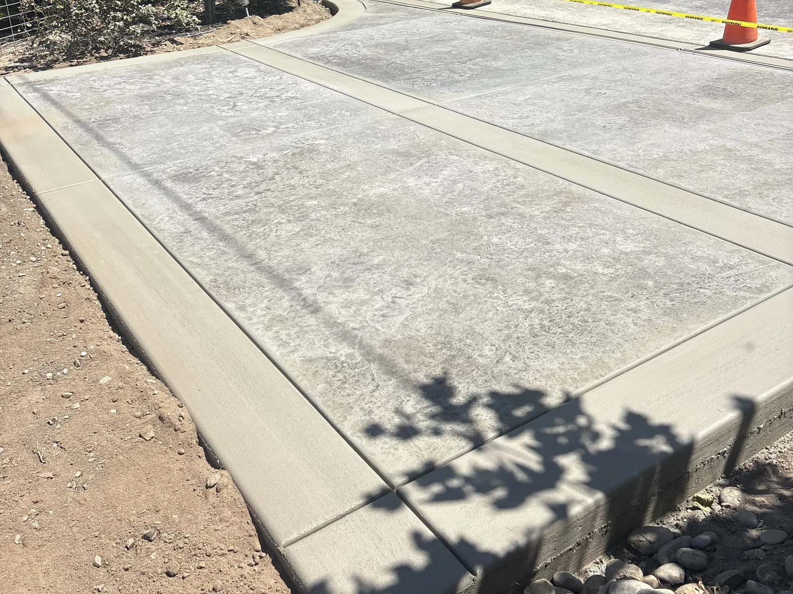 Finished broom-finish concrete driveway pad with a rock border and tree shadows across it at a Modesto-area home