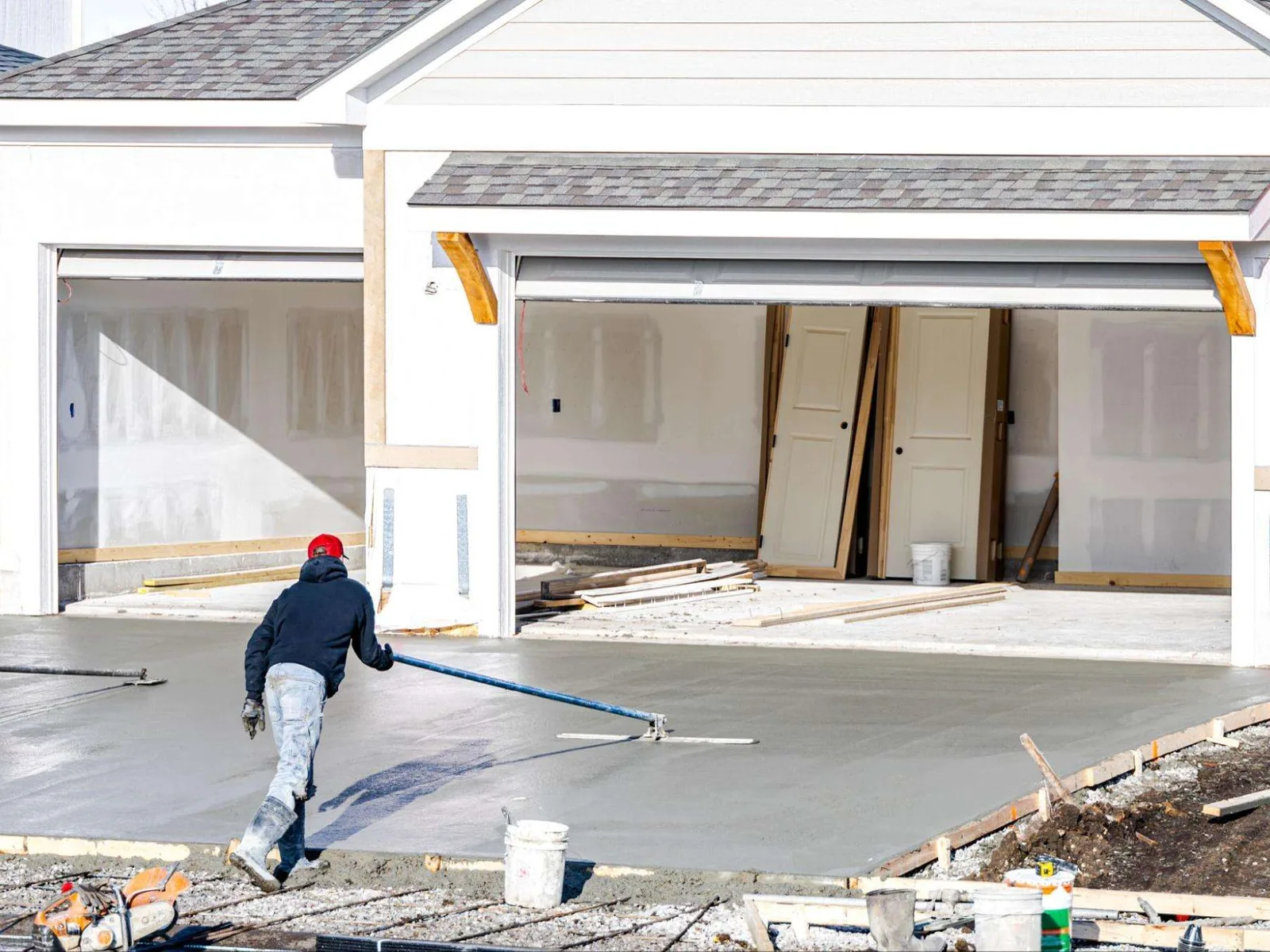 Concrete finisher pulling a bull float across a freshly poured driveway in front of an open three-car garage