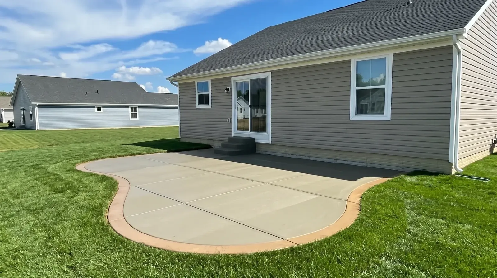 Curved plain concrete patio poured against the back of a house with a green lawn around it