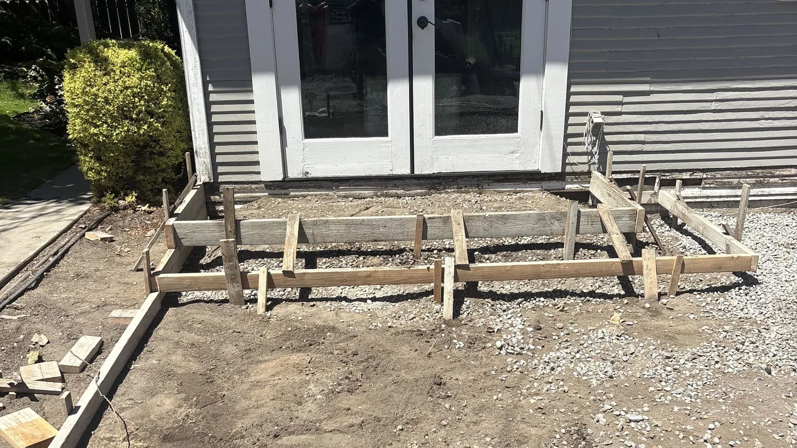 Wood forms and stakes set on a gravel base for a new concrete patio outside a set of white French doors