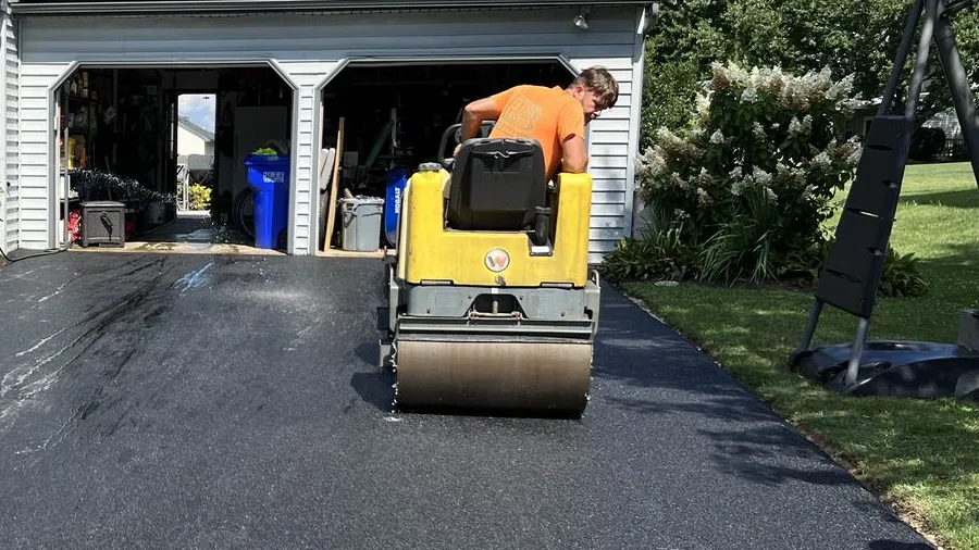 A crew member compacting a new asphalt driveway with a small ride-on roller in front of an open two-car garage