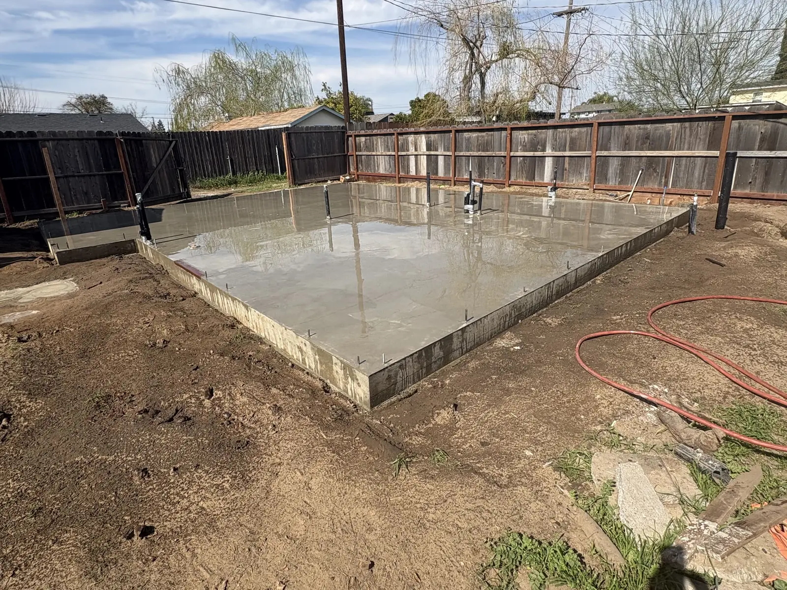 A freshly poured slab foundation in a backyard with anchor bolts and plumbing stubs set in the concrete, a wood fence behind it