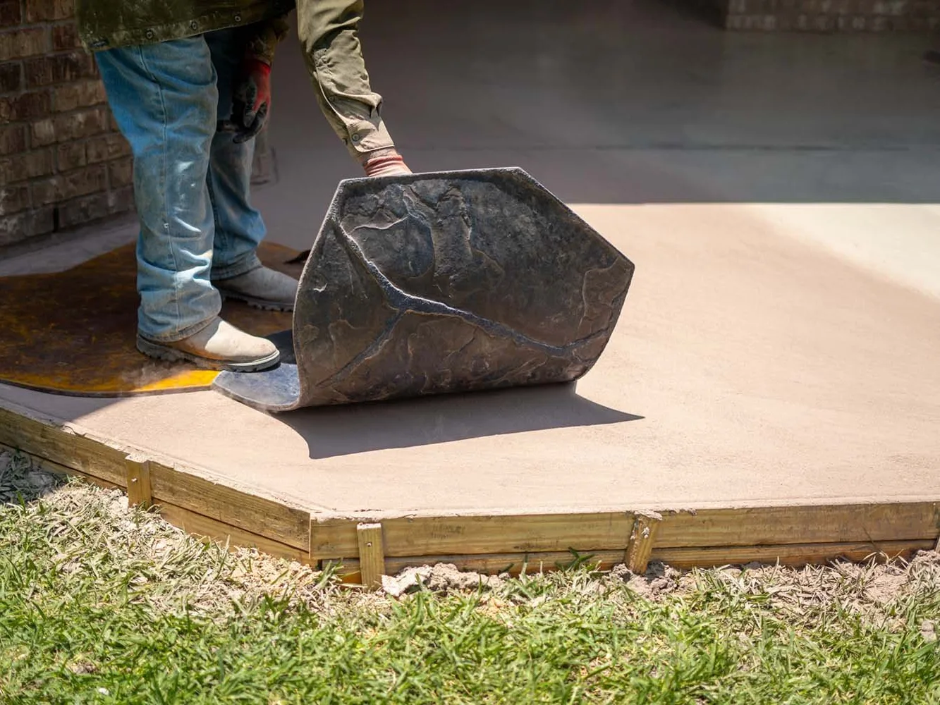 Worker pressing a stone-pattern stamp mat into a freshly poured concrete slab inside wood forms